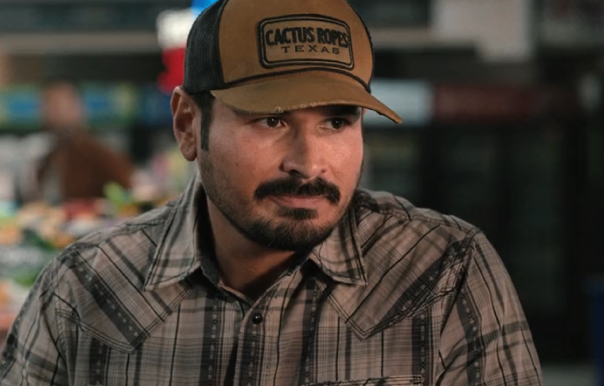 Man wearing a plaid shirt and a cap with "Cactus Ropes Texas" logo, looking thoughtfully in a market setting.