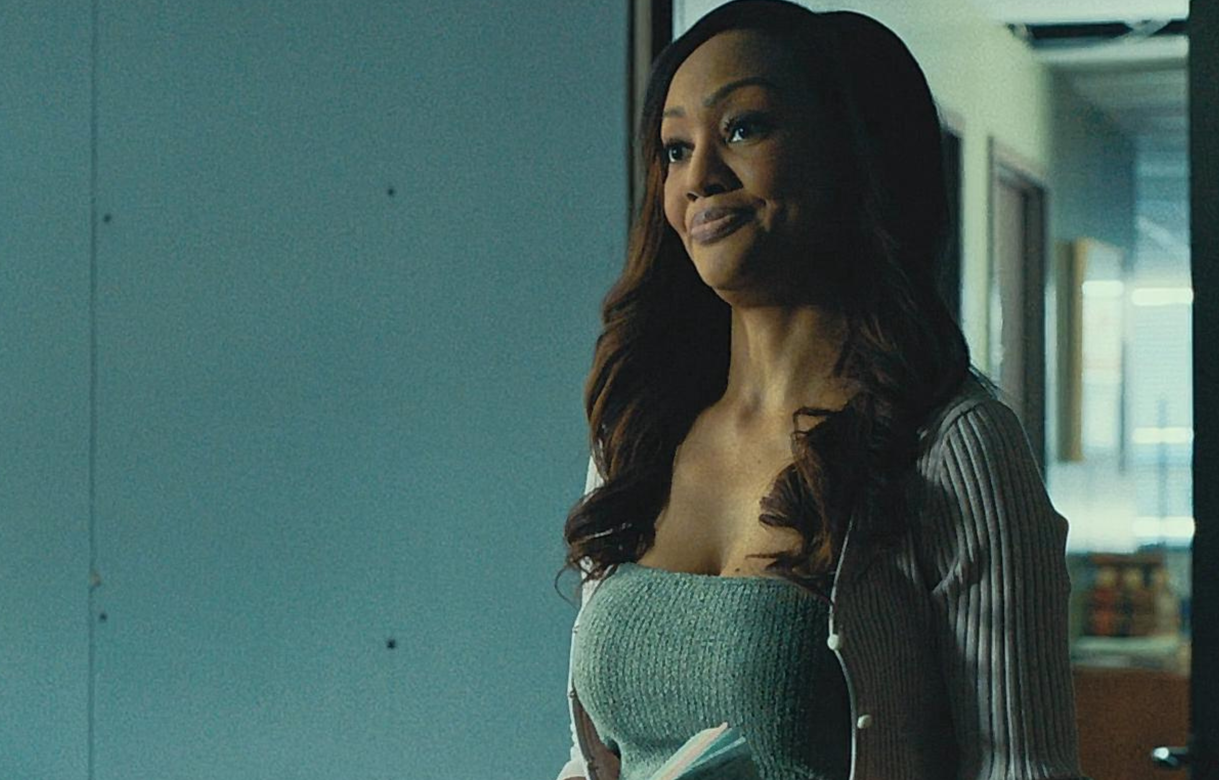 Woman standing in a hallway, smiling, wearing a stylish outfit and holding a notebook.