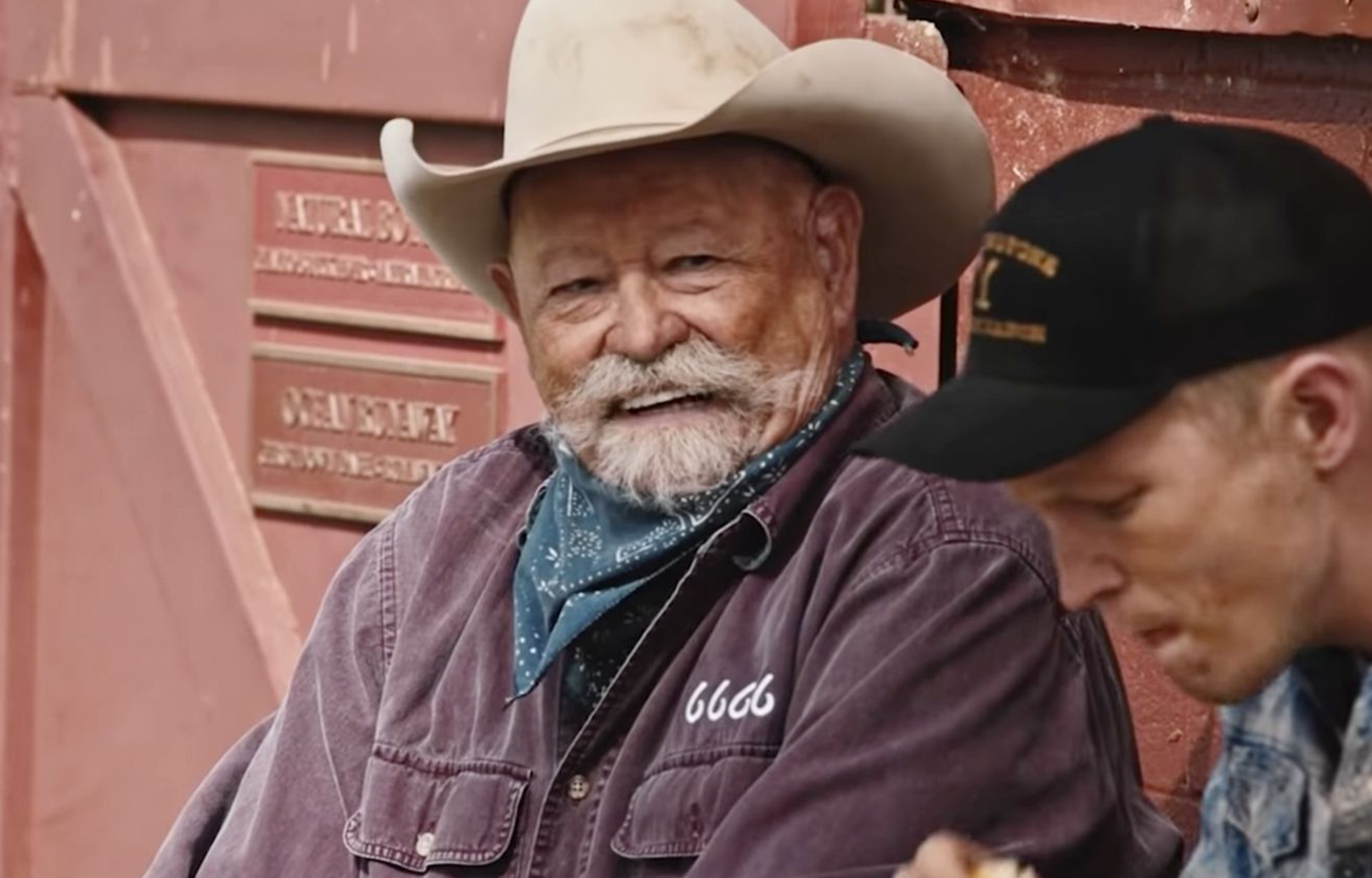 Senior man with a cowboy hat and bandana smiling while sitting next to a younger man eating.