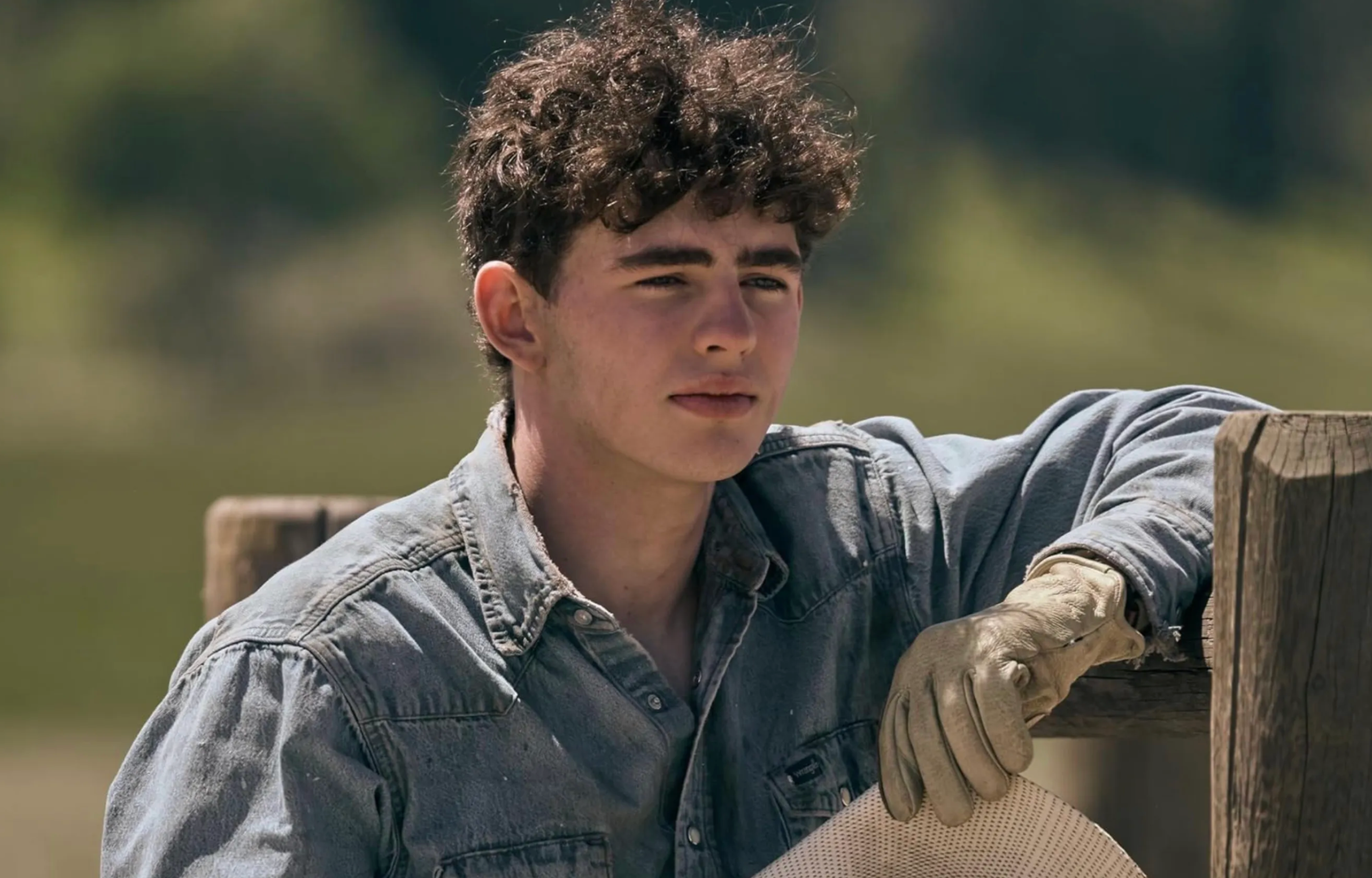 Young man in a denim shirt and gloves leaning on a wooden fence, looking thoughtfully into the distance.
