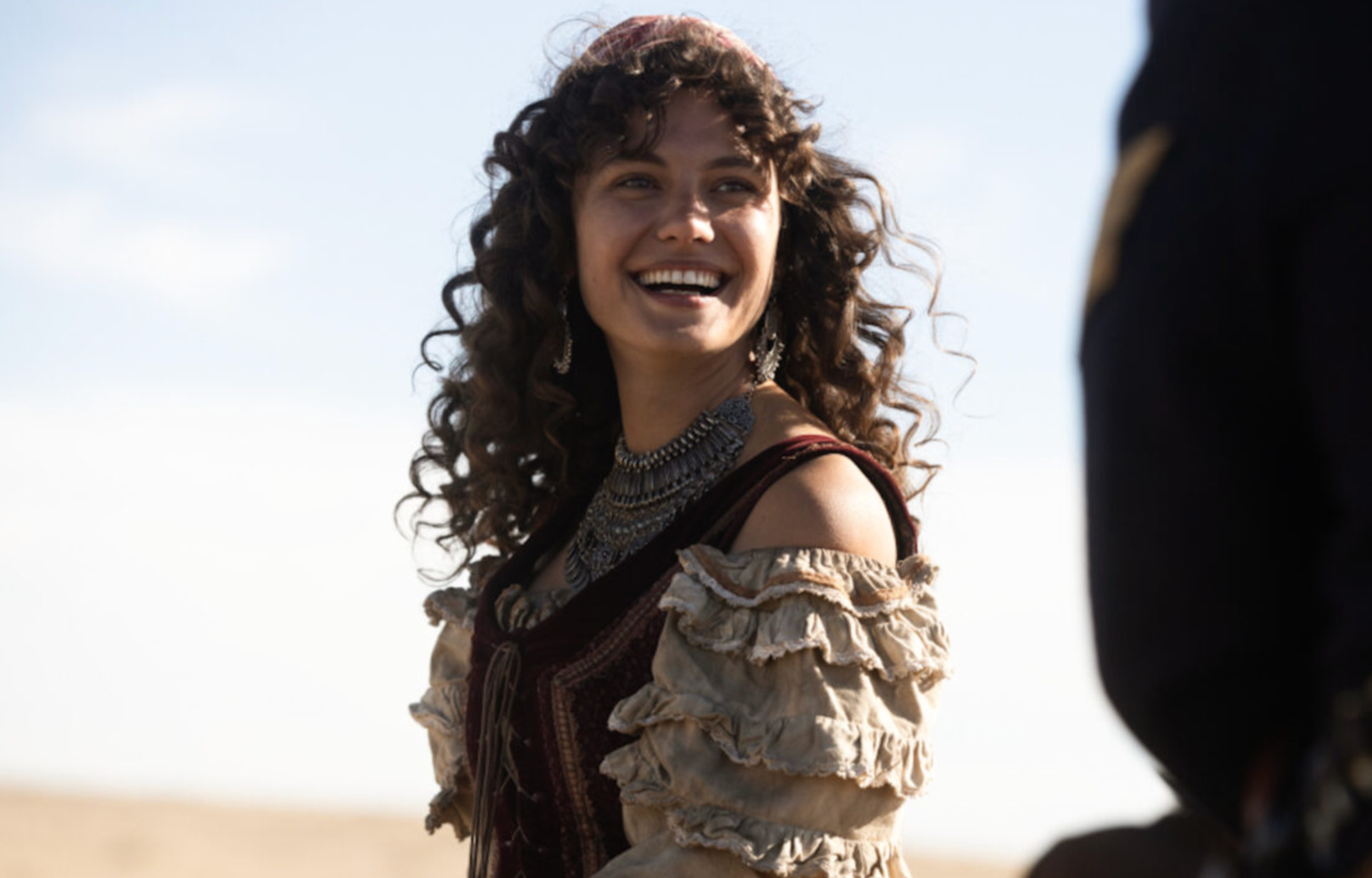 A smiling woman with curly hair wearing a detailed outfit, standing outdoors against a bright sky.