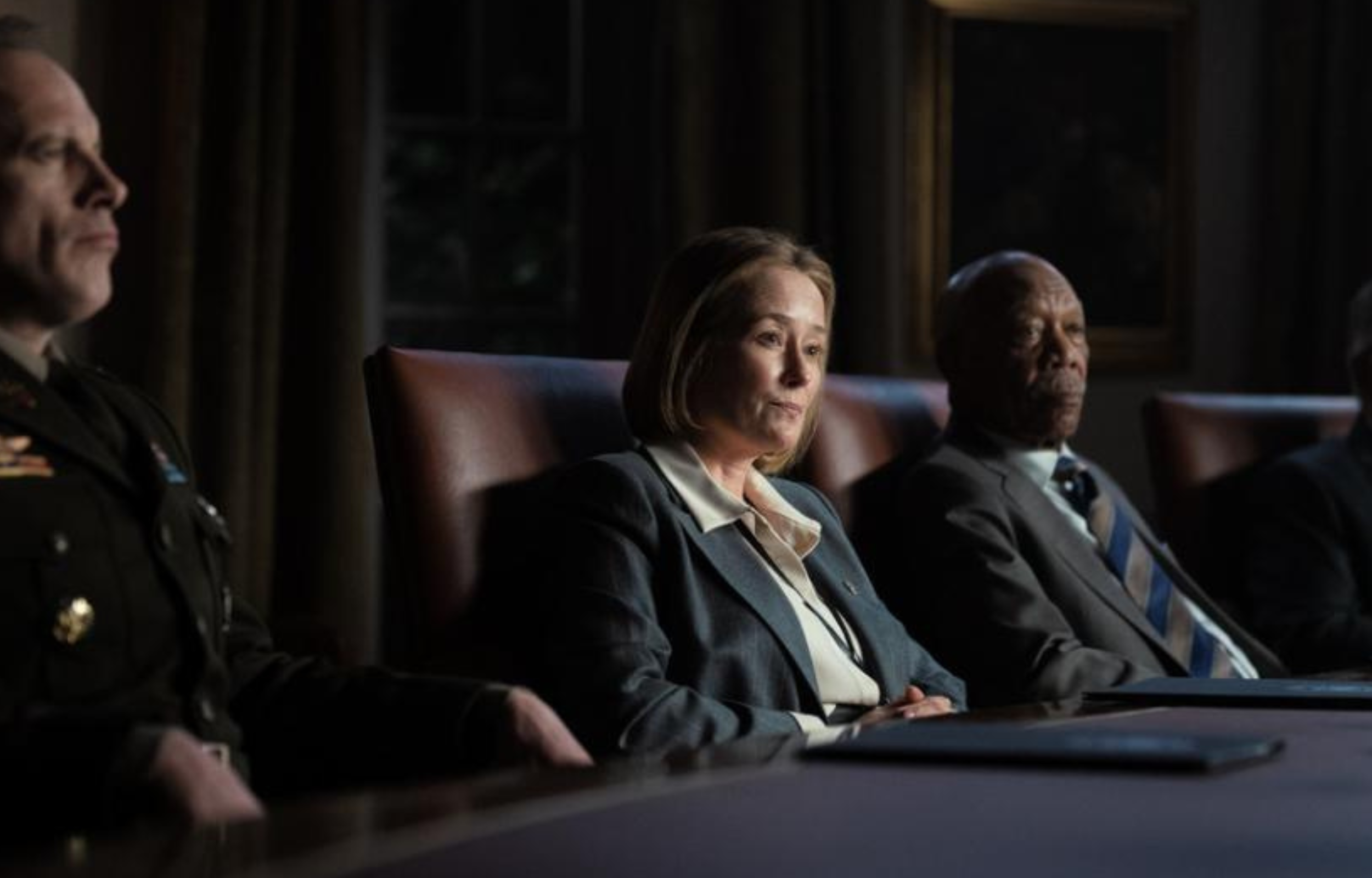 Four people in business attire seated at a long table in a dimly lit room, discussing serious matters.