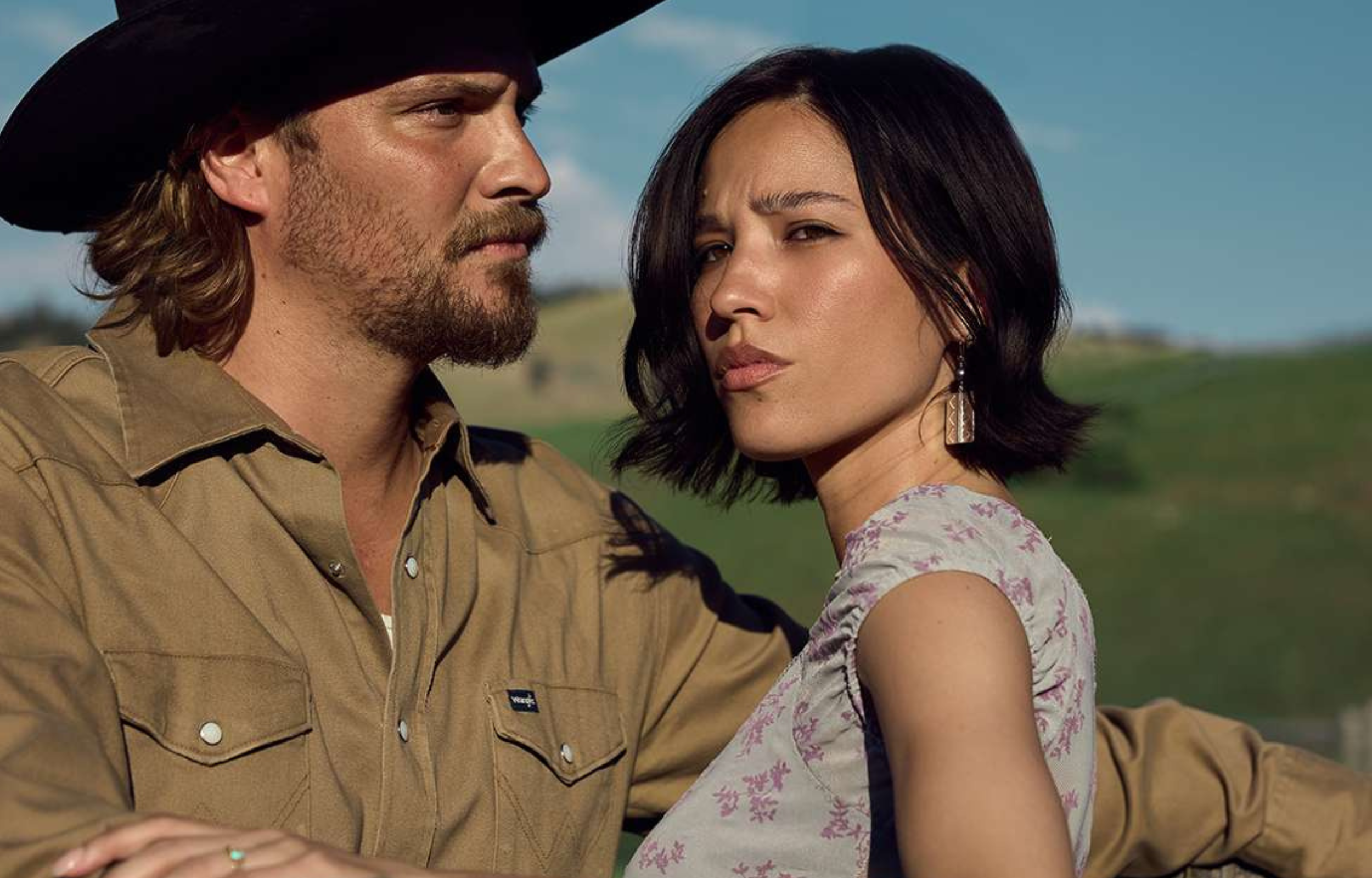 A man and woman outdoors, both looking serious, with a rural landscape in the background.