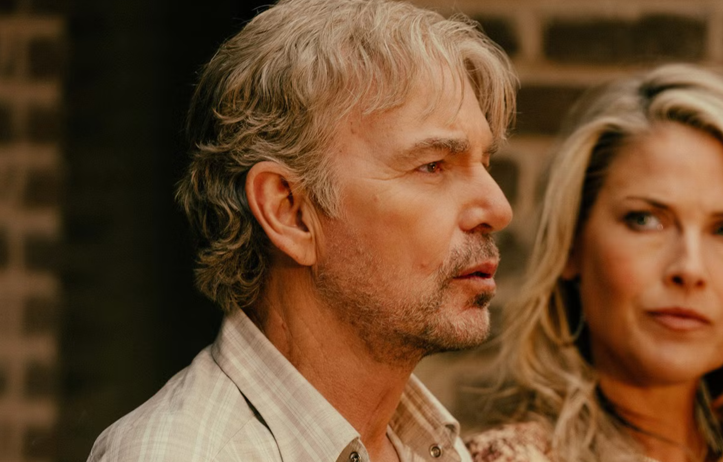 Two people in profile, with a focus on a man with silver hair and a woman with blonde hair, against a brick backdrop.