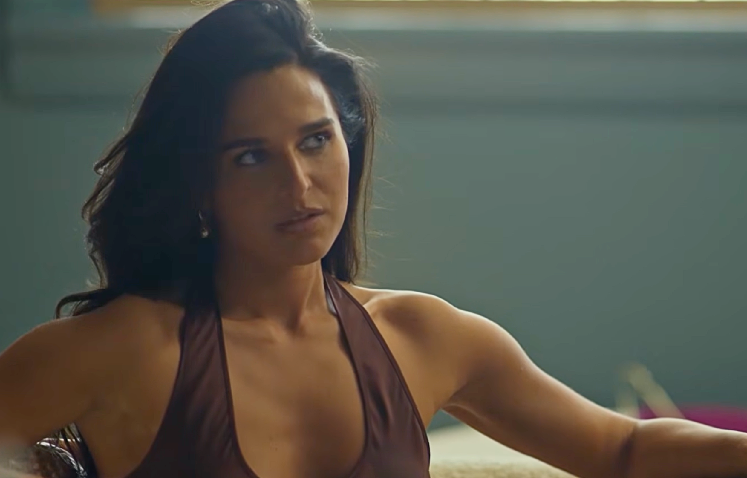Woman with long hair sitting and looking thoughtfully, wearing a brown sleeveless top.