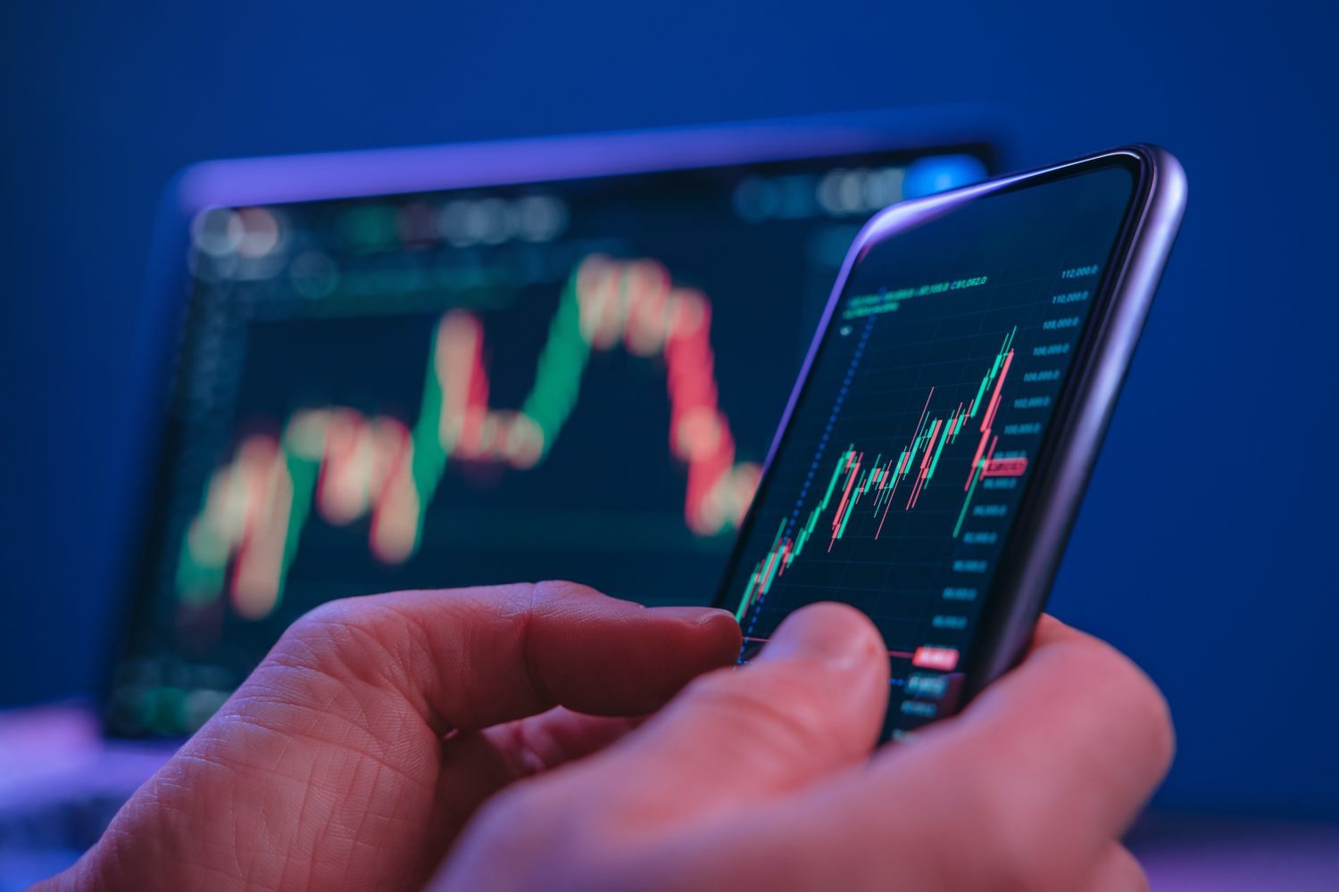 Close-up of hands holding smartphone displaying a stock market candlestick chart, with laptop screen in background. Trader analyzing financial charts on a phone late at night