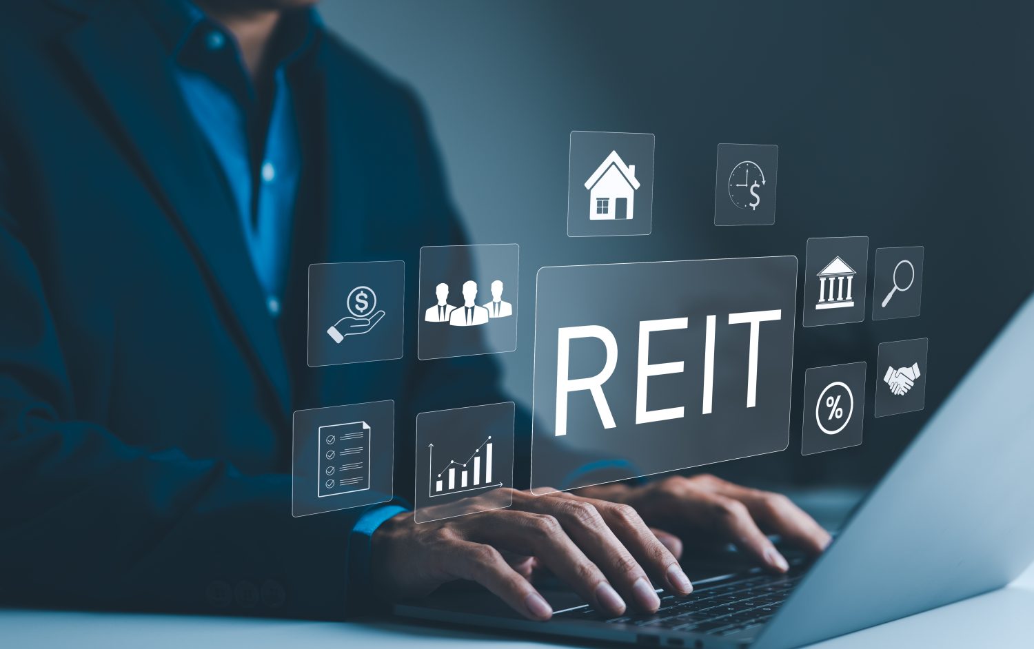 A person in a dark suit types on a silver laptop, their hands visible on the keyboard. Above the laptop, a dark blue-tinted holographic interface displays numerous white outline icons representing financial and real estate concepts, such as a house, dollar signs, a bar graph, a handshake, and a percentage sign. The word "REIT" is prominently centered in large white text among these glowing icons. The background is a blurred dark office setting.