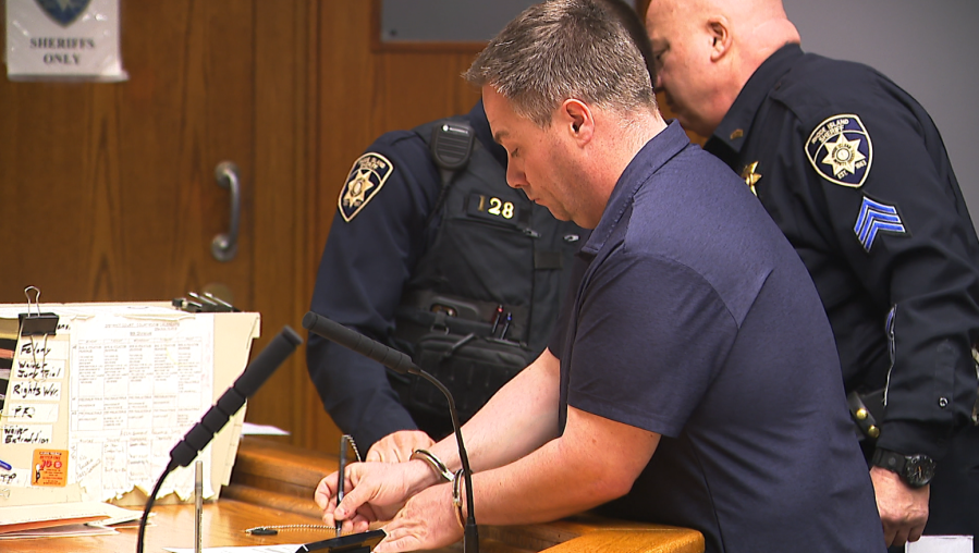 Nicholas LeBlanc stands at a courtroom lectern in handcuffs with Rhode Island sheriff's officers behind him.