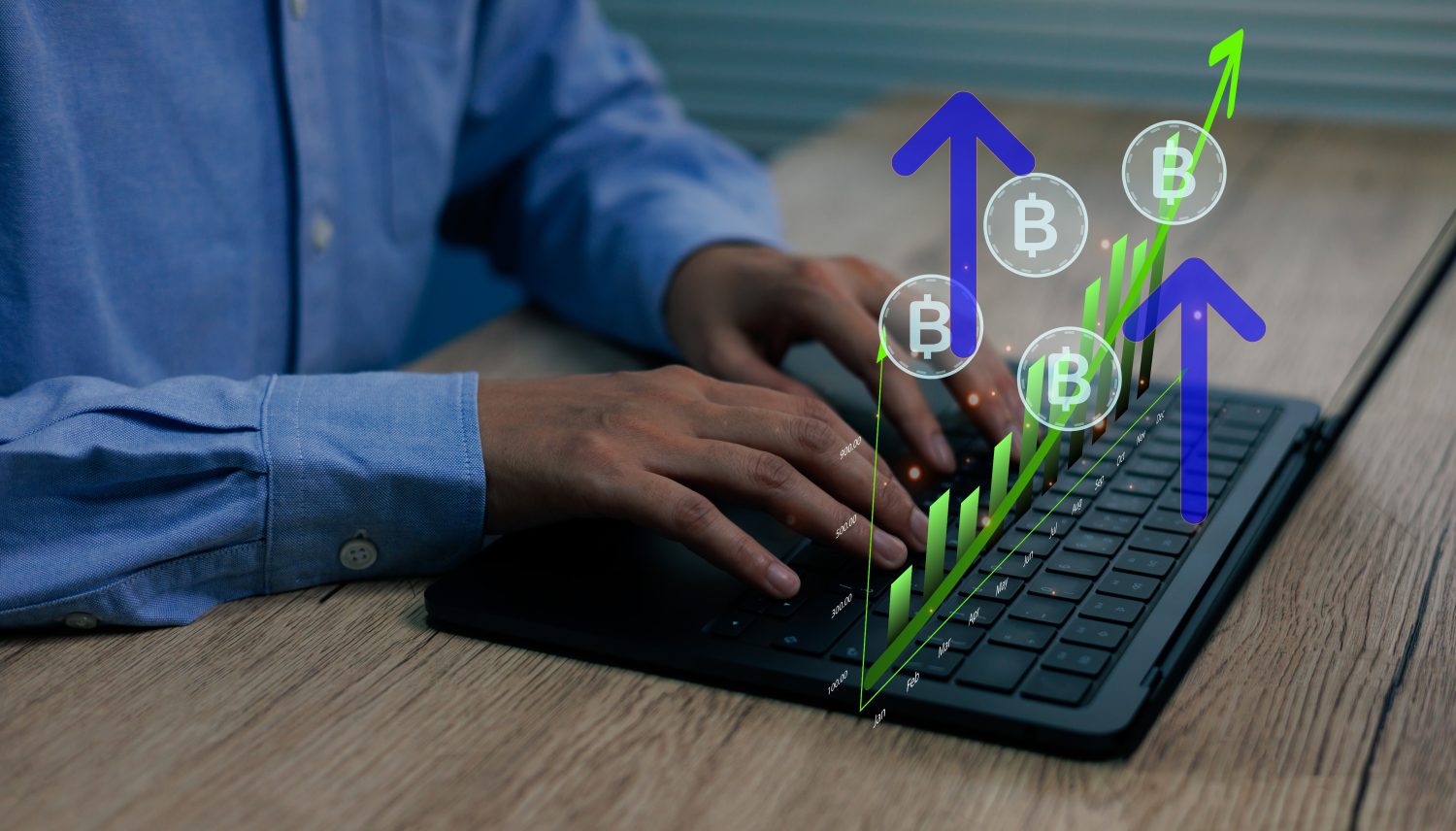 A close-up shot of a person's hands typing on a black laptop keyboard. Overlayed on the screen and extending above are digital graphics showing an upward-trending green bar chart with monetary values and months, several blue upward-pointing arrows, and multiple circular icons with a white 'B' symbol representing Bitcoin. The person is wearing a blue long-sleeved shirt, and the laptop rests on a wooden desk.