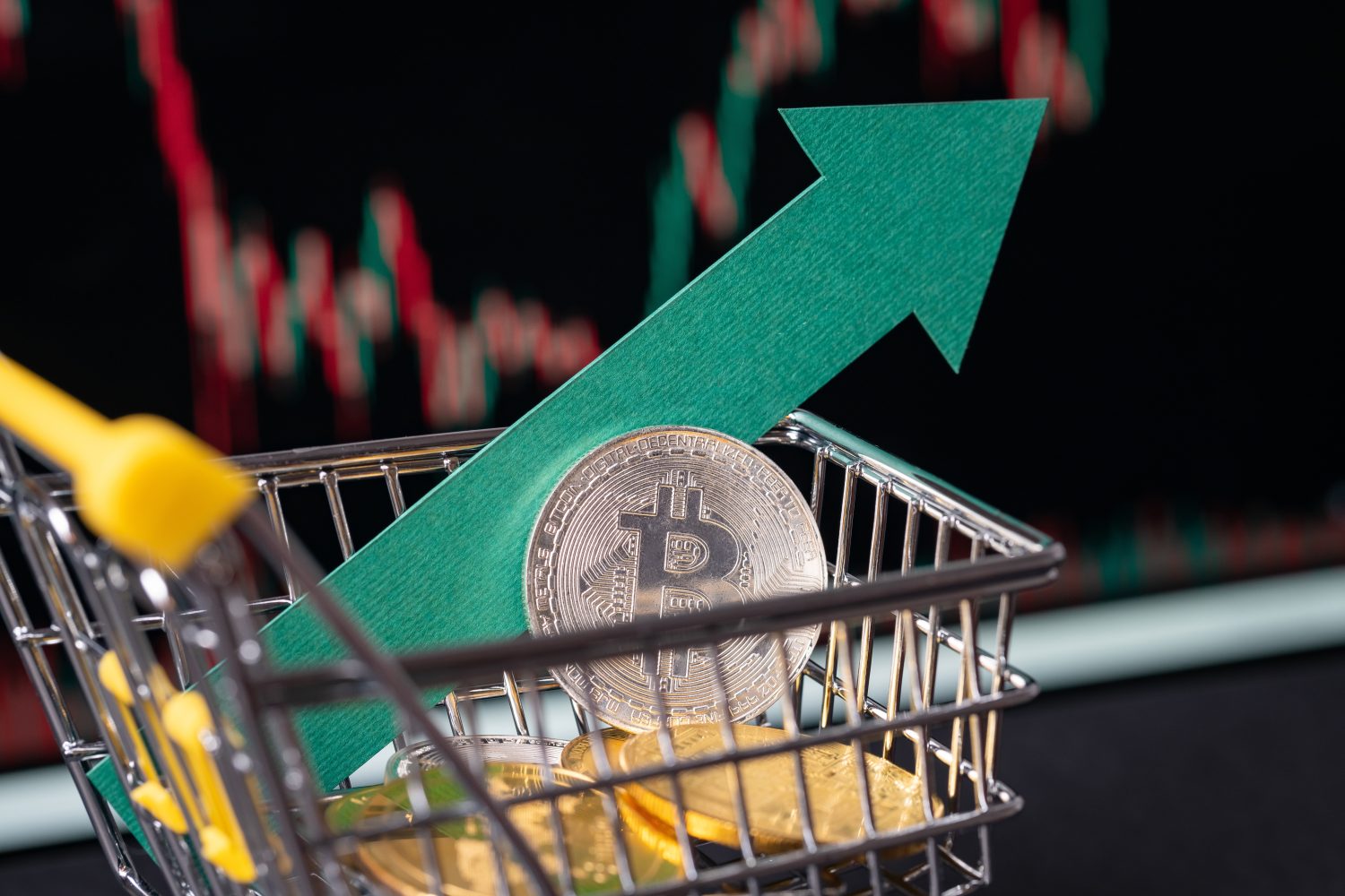 A close-up shot of a miniature metal shopping cart. Inside, a silver Bitcoin coin stands prominently next to several golden coins. A vibrant green arrow points diagonally upwards from the bottom left, passing through the cart. The background is softly blurred with abstract red and green patterns, resembling a financial stock chart.