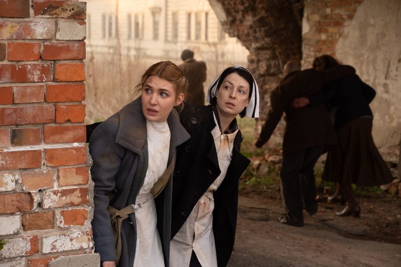 Two nurses look scared as they hide behind a brick wall
