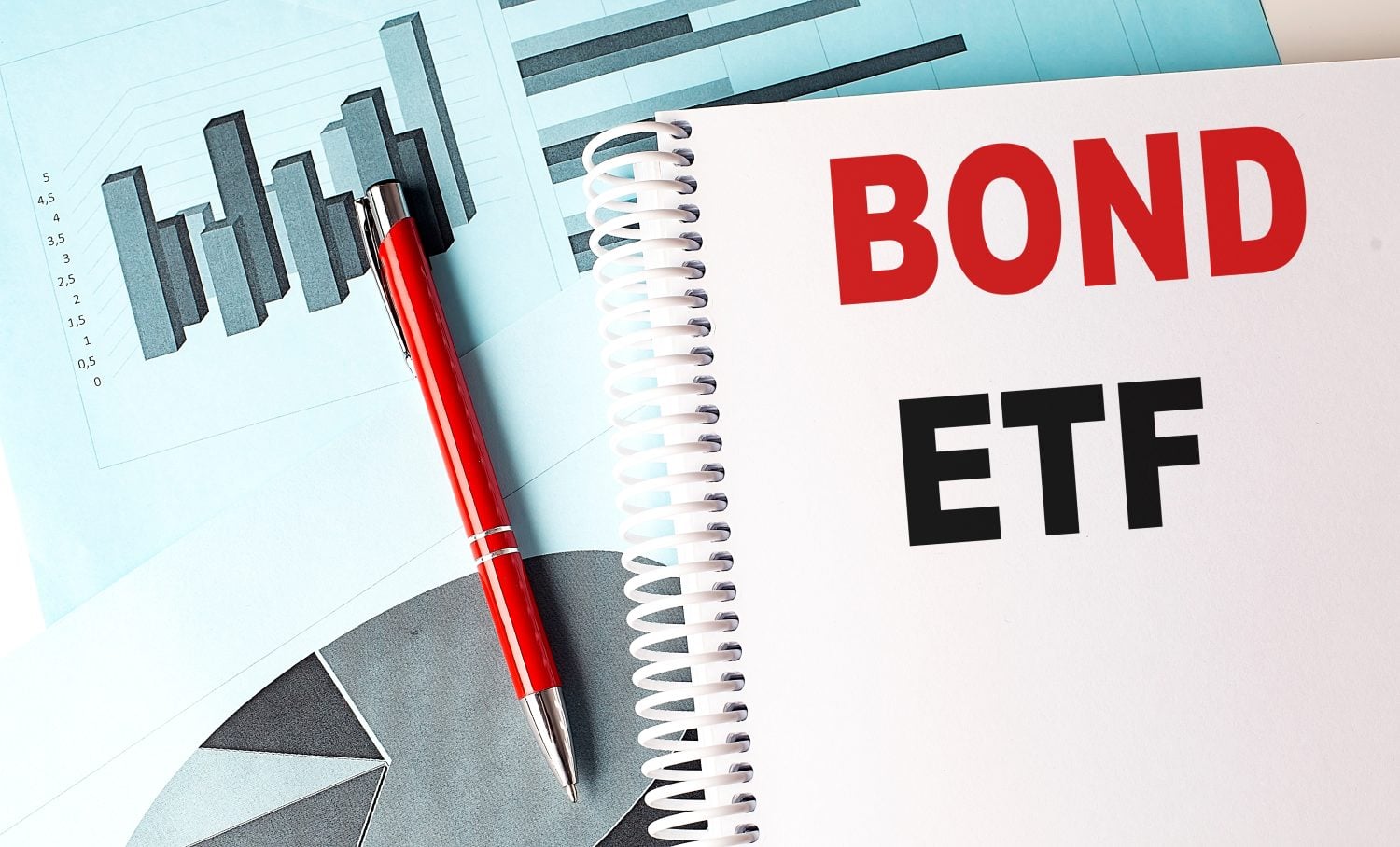 A red pen rests on an open, spiral-bound notebook. The notebook page features the word 'BOND' in large red letters and 'ETF' in large black letters below it. In the background, a light blue financial document with a grey bar graph and a partial pie chart is visible.