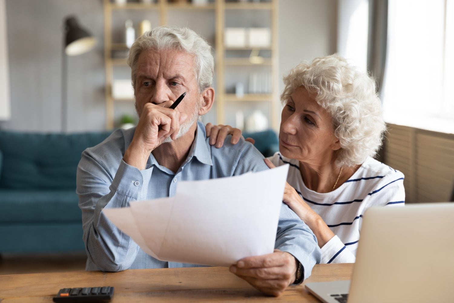 A distressed older man with gray hair and a beard holds a pen and papers, resting his chin on his hand, looking down. An older woman with curly gray hair gently places her hand on his shoulder, looking at him with concern. They are seated at a wooden table with a calculator and a silver laptop partially visible, in what appears to be a home office or living room.