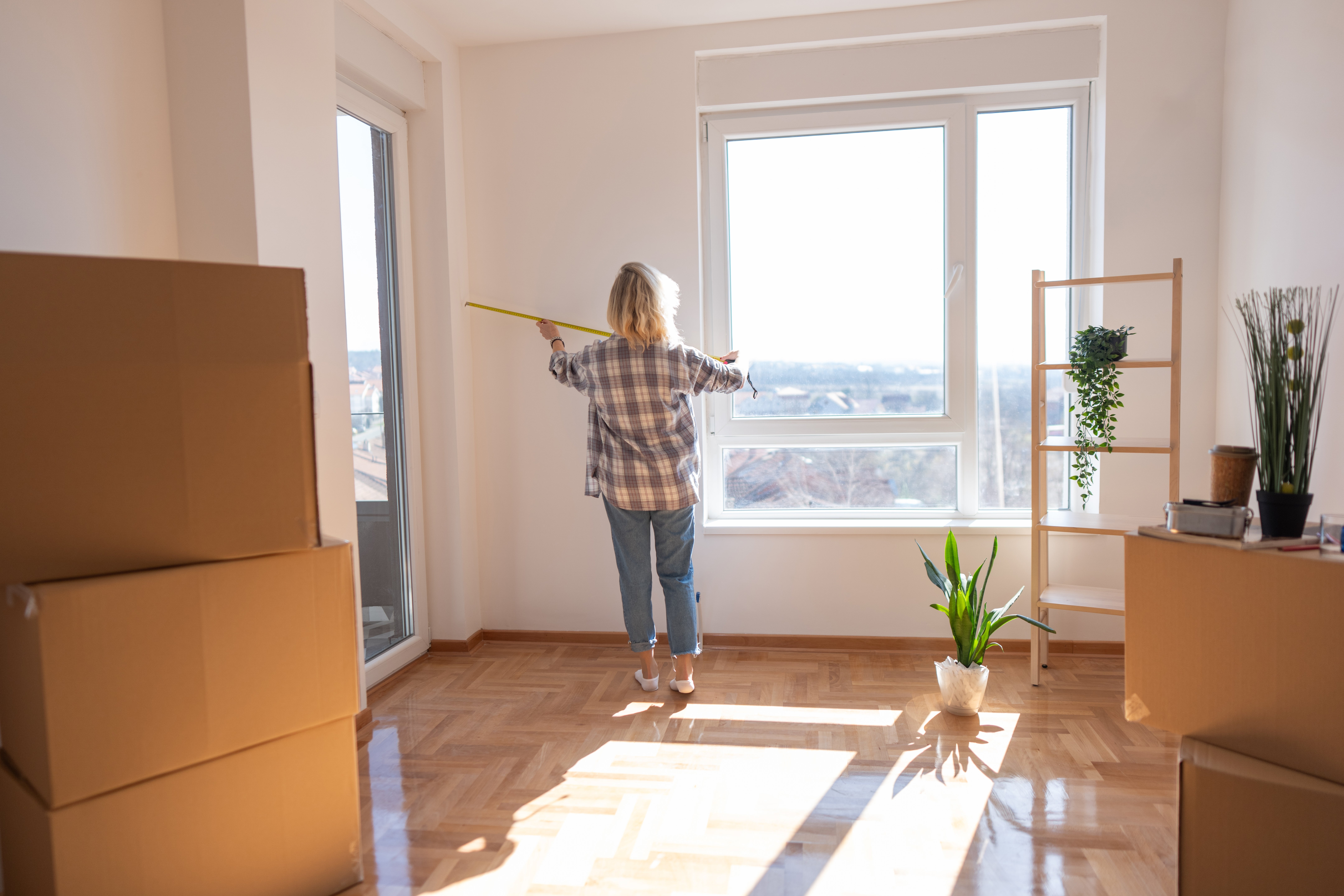 Person measuring window in bright, unfurnished room with packed boxes, indicating a move or property renovation
