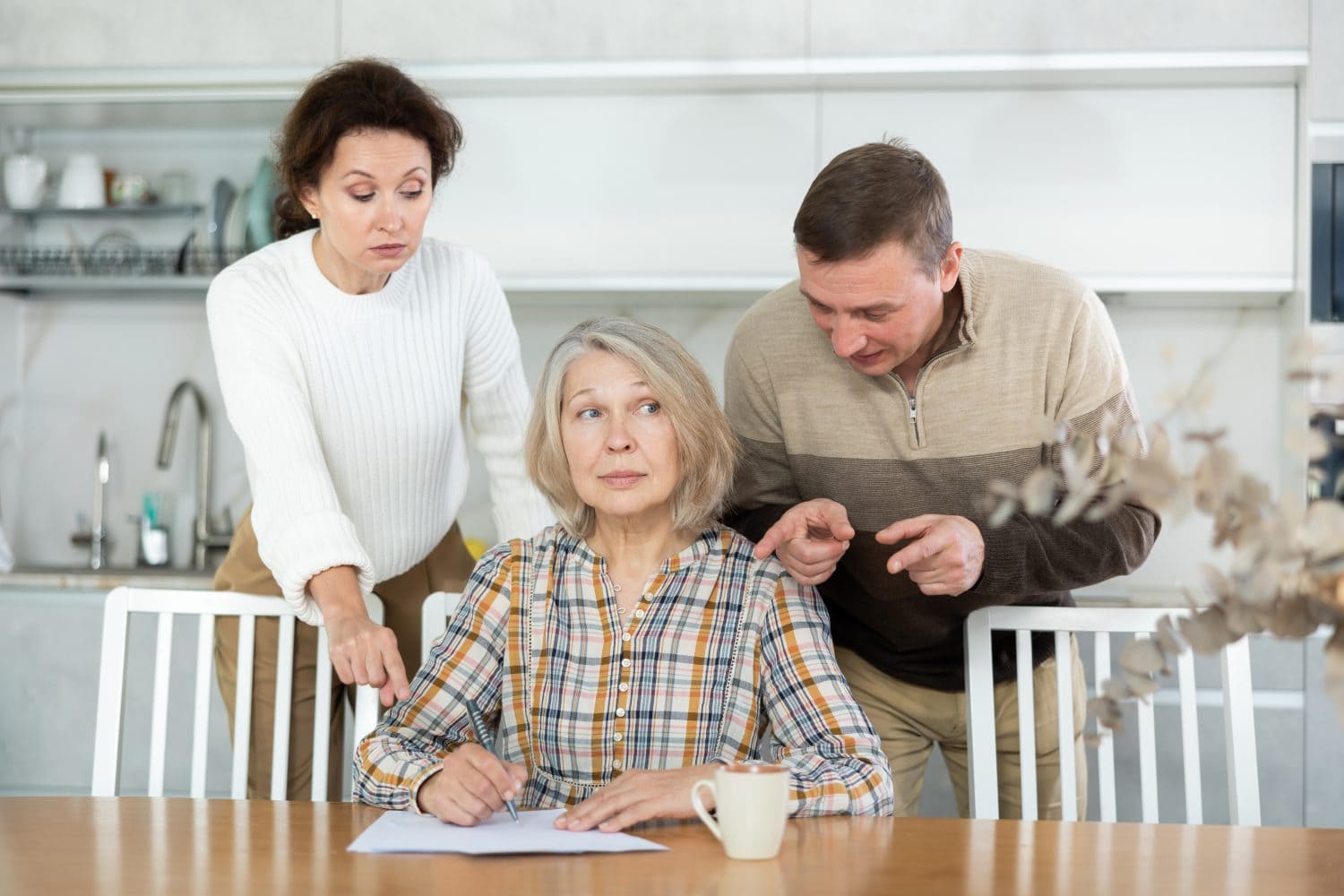 An older woman with gray hair, wearing a plaid shirt, sits at a light wooden table, holding a pen over a document. She looks to her right with a concerned expression. Behind her, a younger woman in a white sweater and a man in a beige and brown striped sweater lean in, both looking at the document and pointing. A white coffee mug is on the table. The background features a modern kitchen with white cabinets and a sink.
