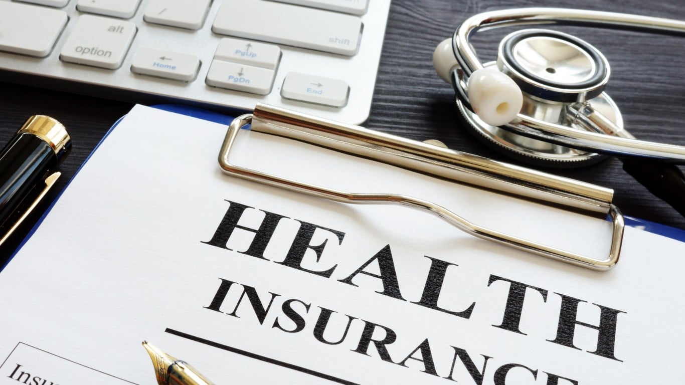 A close-up of a document titled 'HEALTH INSURANCE' on a blue clipboard. A black and gold fountain pen lies to the left of the document. A silver stethoscope is positioned on the right. In the blurred background, a silver computer keyboard is partially visible. The overall scene is set on a dark wooden surface.