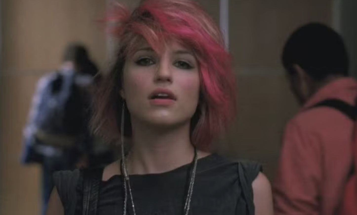 Person with short, styled hair, wearing a sleeveless top and necklace, looking forward. Background shows people in casual attire