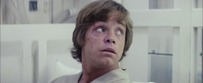 Luke, having short hair looking surprised while seated, wearing a light-colored robe, in a medical or sci-fi setting