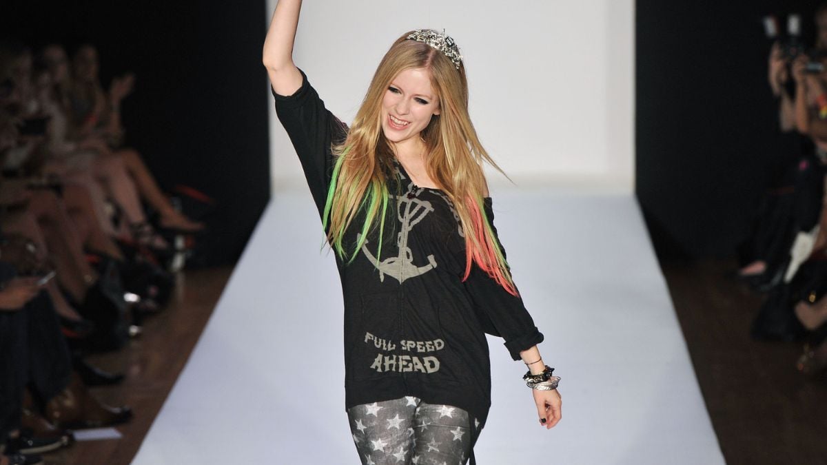 A woman with colorful hair walks a runway, smiling and raising an arm. She wears a black shirt with “FULL SPEED AHEAD” text, star-patterned leggings, and a crown headband.