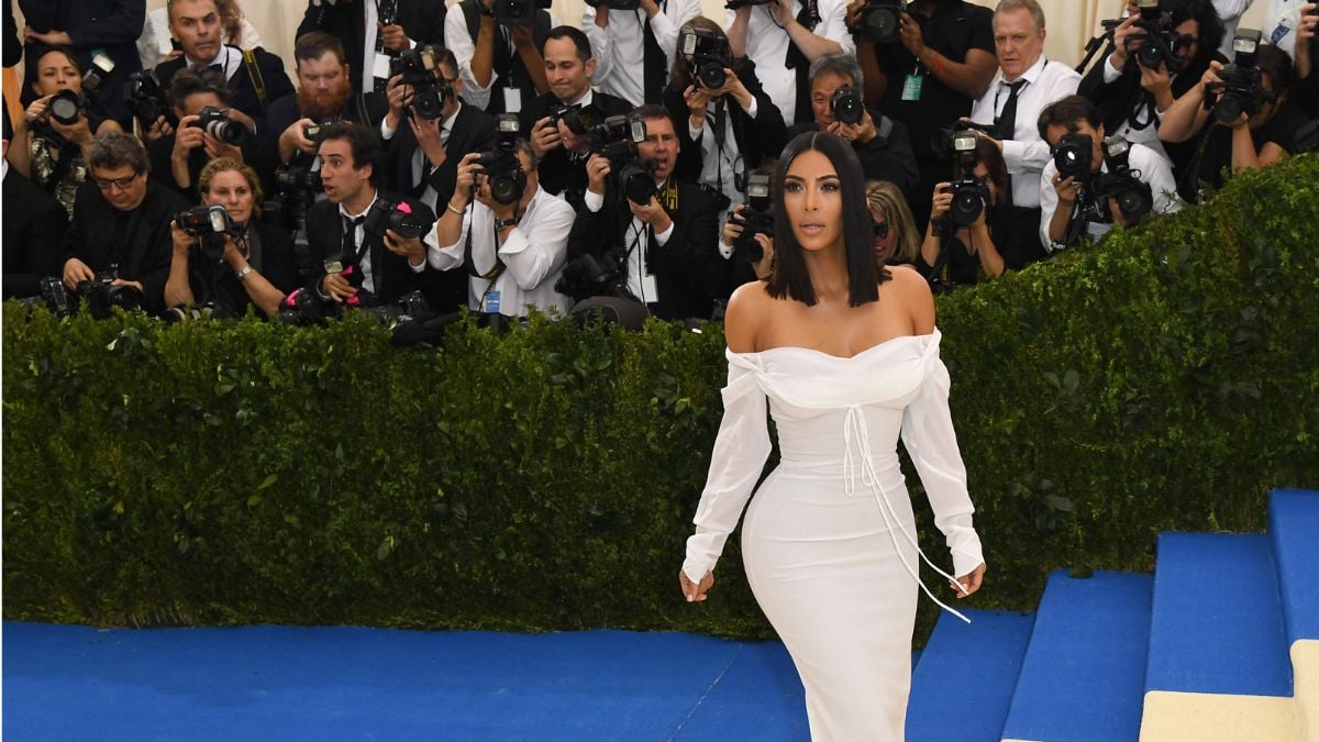 A woman in an off-shoulder white dress poses on blue stairs at an event, surrounded by photographers and lush green foliage in the background.