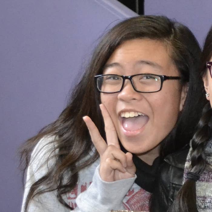 Person smiling and holding a peace sign, wearing glasses and a casual outfit, shoulder-length hair visible