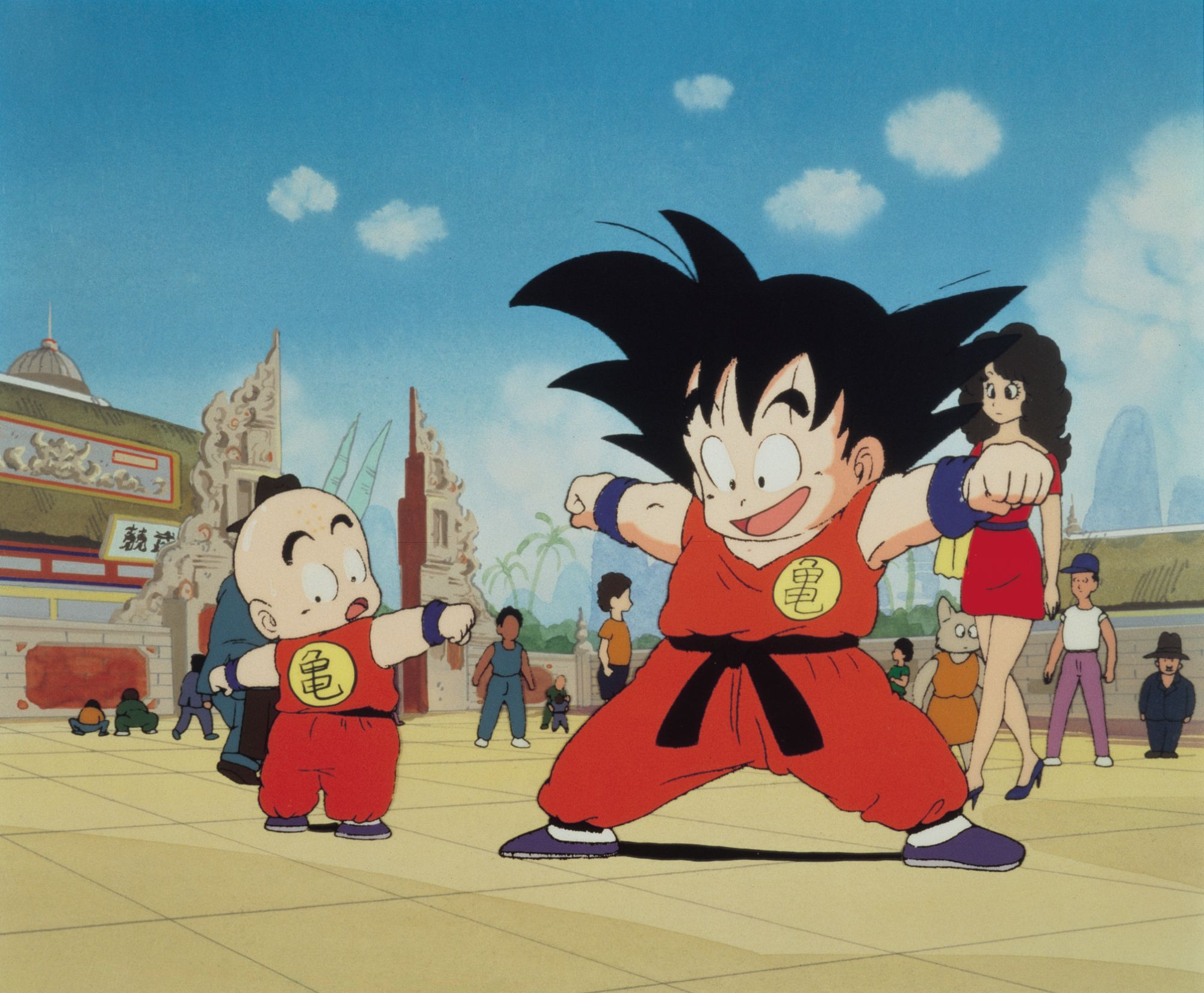 Anime characters Goku and Krillin in martial arts stances, wearing traditional orange outfits in an open courtyard setting