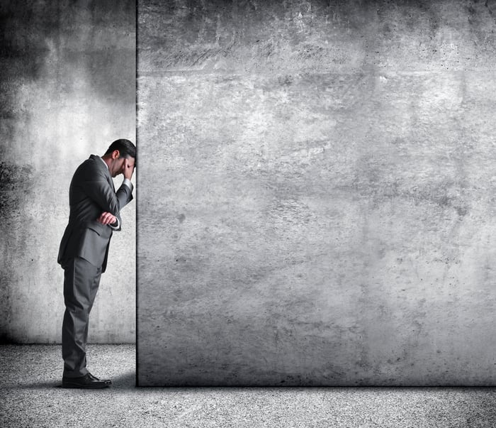 A worried investor rests his head against a gray wall.