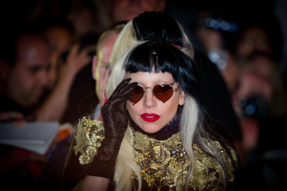 Lady Gaga wearing heart-shaped sunglasses, brown gloves, and an ornate gold-embellished top while surrounded by a crowd.