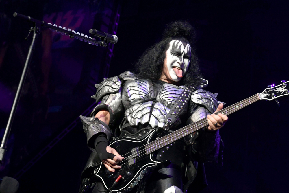 Gene Simmons of the band KISS wearing demon makeup and spiked armor while playing a black bass guitar on stage with his tongue out.
