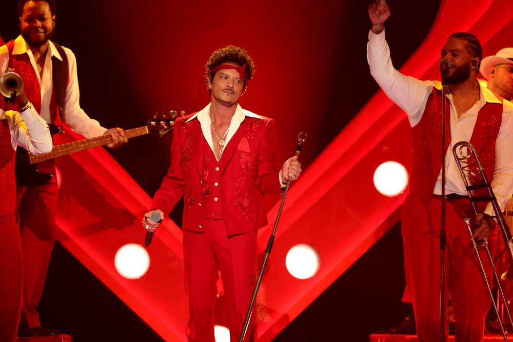 Bruno Mars performing on stage in a red suit with a headband, flanked by band members in matching red outfits.