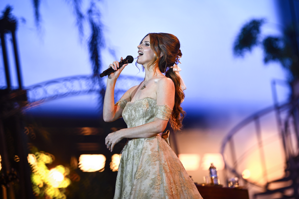 Lana Del Rey singing into a microphone on stage while wearing a tan lace gown.