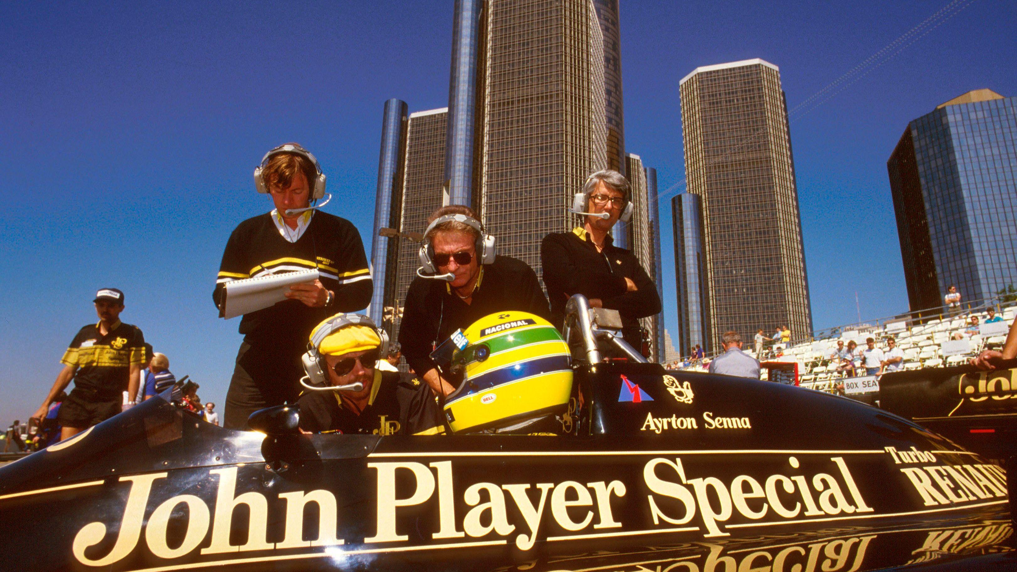 Ayrton Senna sat in his black and gold Lotus, speaking to Lotus team members, with high-rise buildings in the background before the 1986 Detroit Grand Prix 