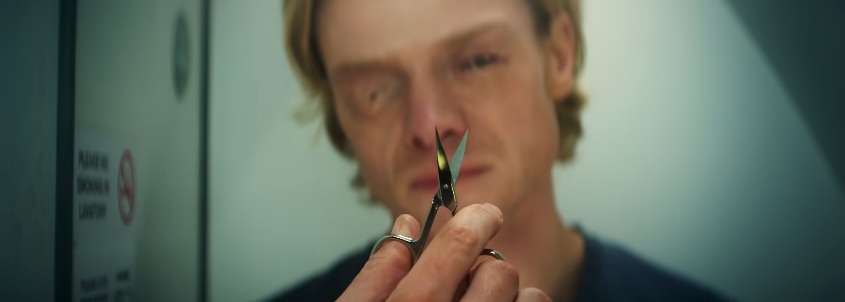 A close-up of a person holding a pair of scissors with a serious expression in a dimly lit bathroom setting.