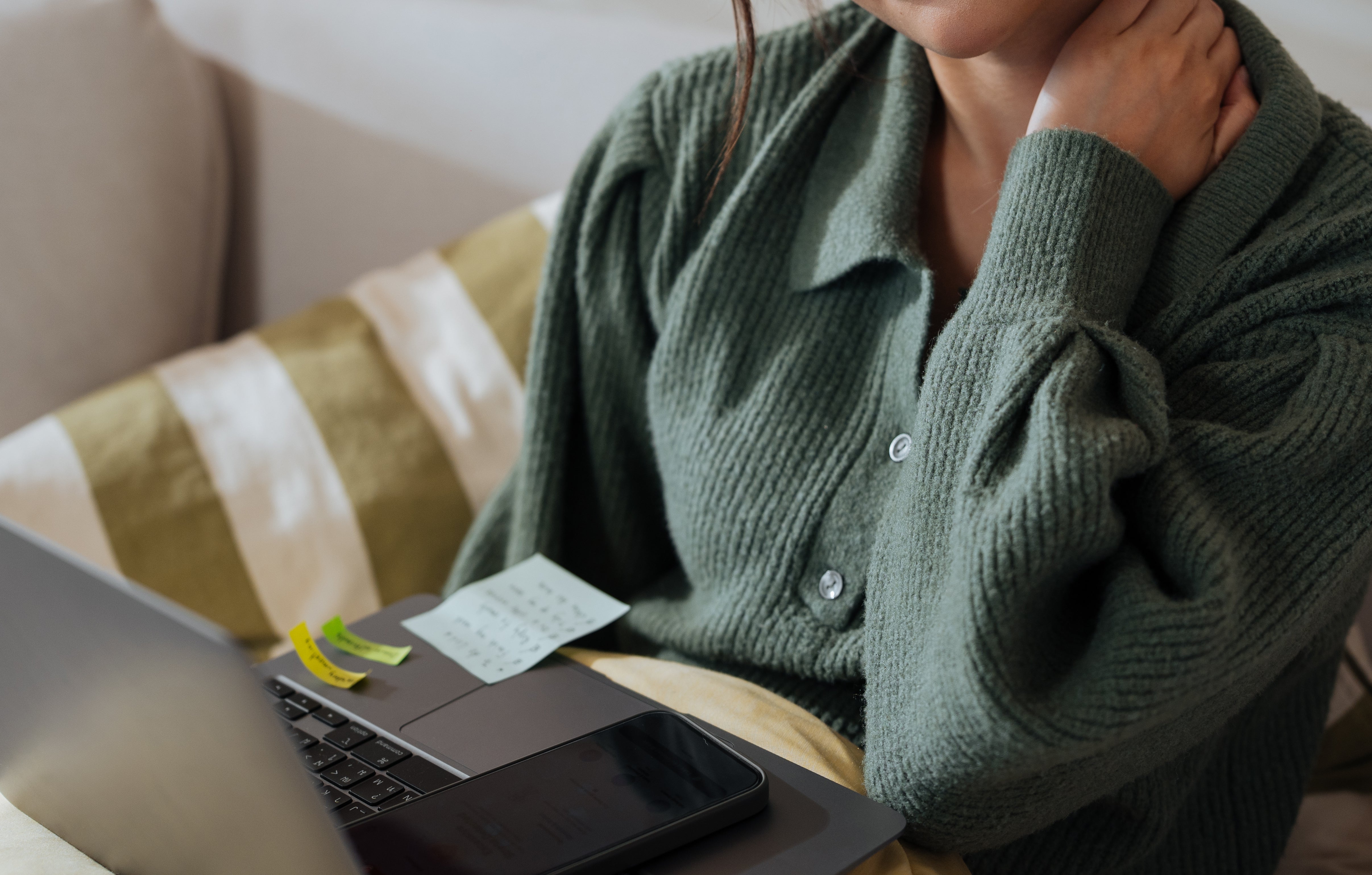 Person sitting on sofa with laptop, rubbing neck, looking tired or tense. Sticky notes and phone are on the laptop