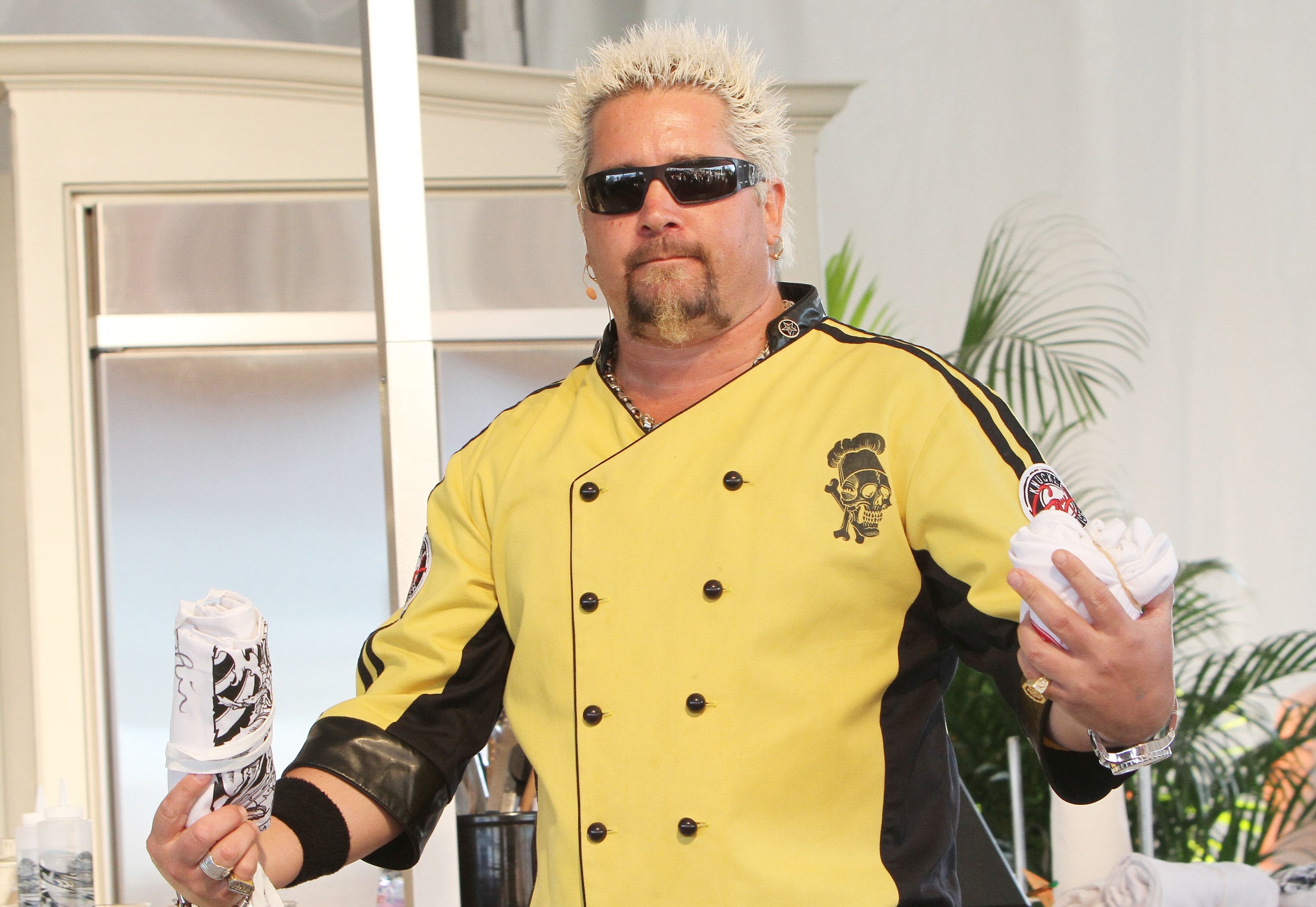 Guy Fieri wearing a yellow and black chef's jacket with sunglasses, holding objects with both hands, standing on a stage
