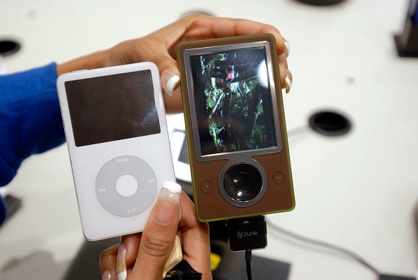 Person holding an iPod and a Zune side by side, comparing two popular media players from the 2000s