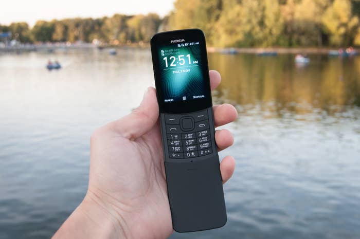 Hand holding a retro style Nokia flip phone, showing the time 00:51, by a lake with blurred trees and boats in the background